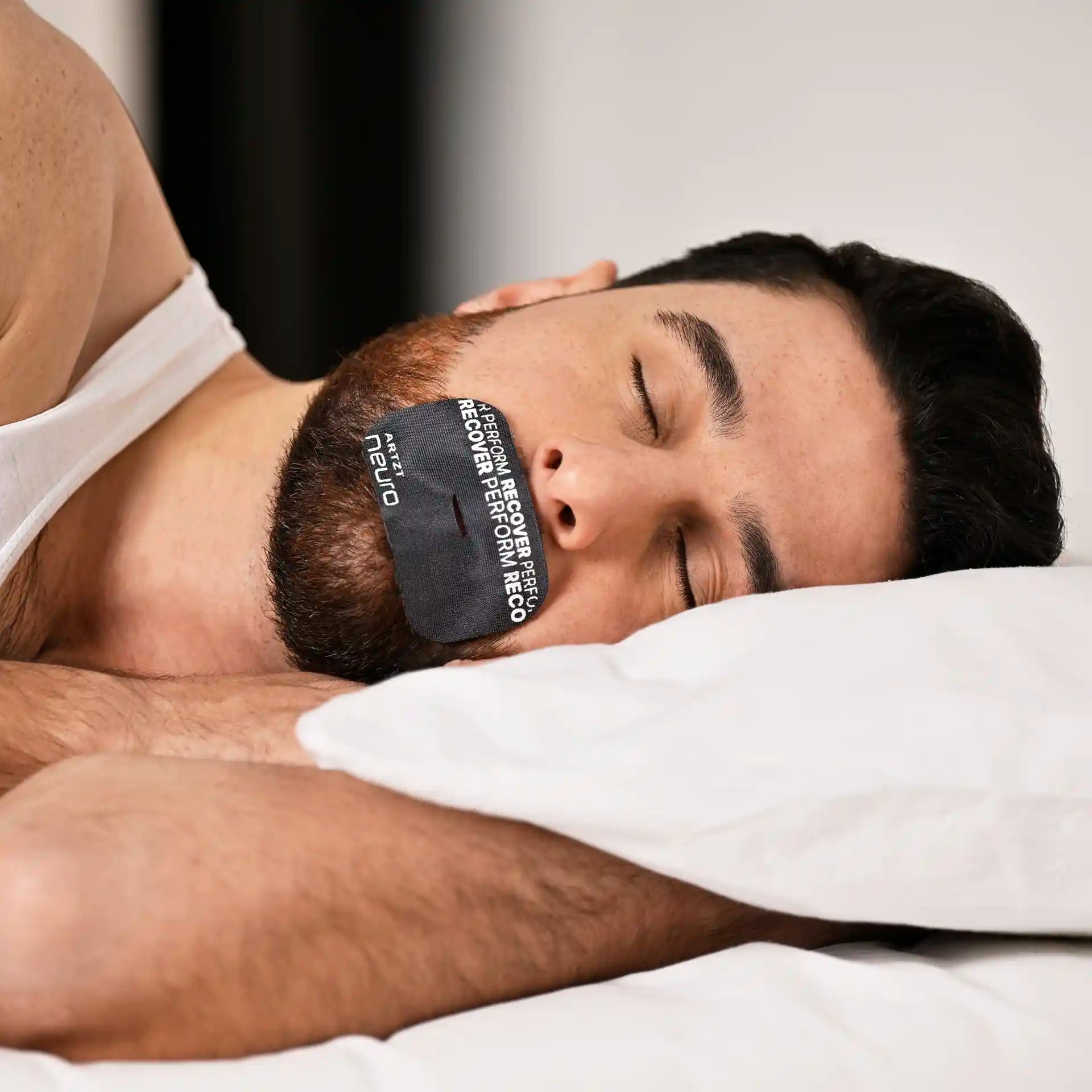 Eine Mann liegt mit geschlossenen Augen auf der Seite in einem Bett mit weißen Laken und trägt auf dem Mund ein Midnight-farbenes Mouth Tape Recovery #farbe_midnight