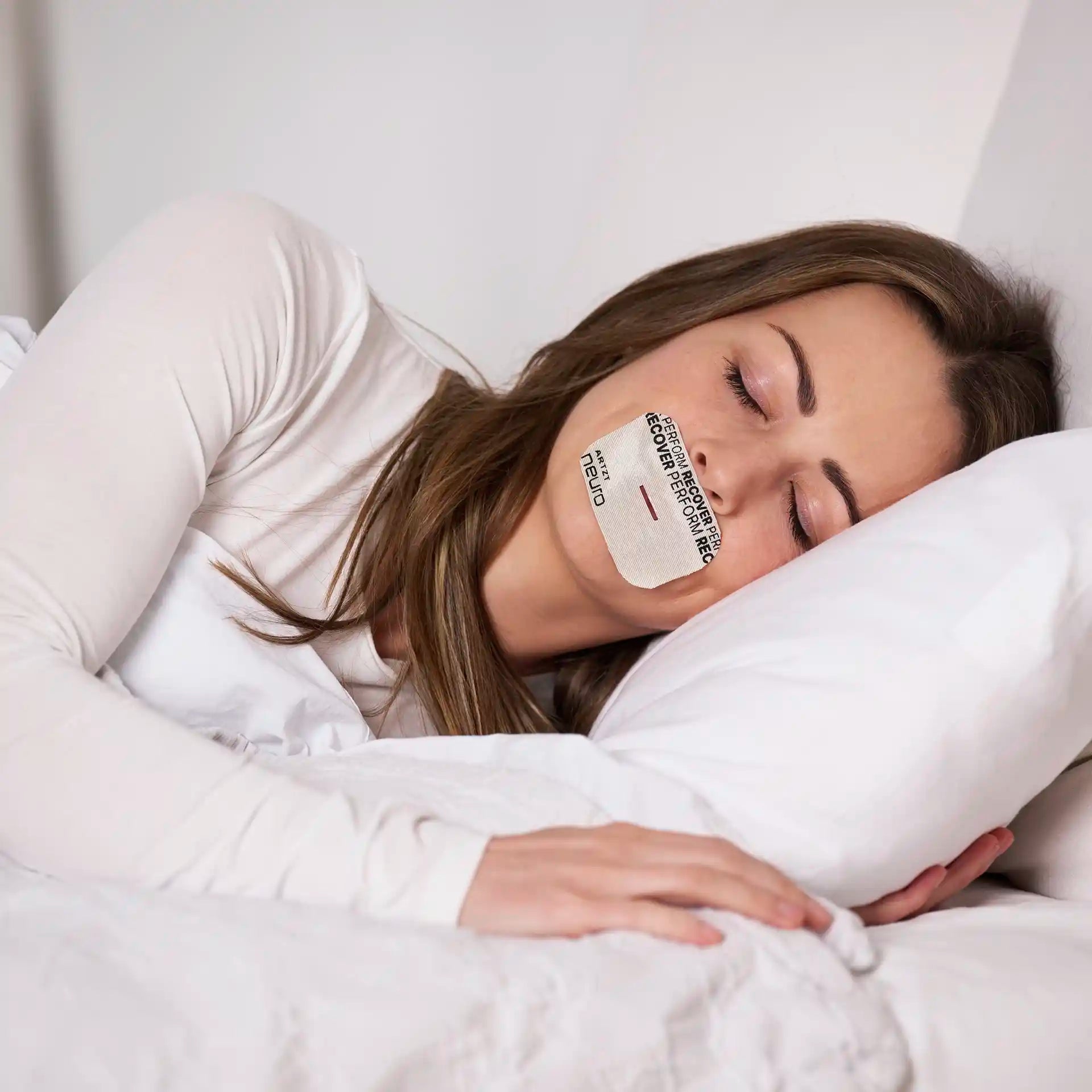 Eine Frau liegt mit geschlossenen Augen auf der Seite in einem Bett mit weißen Laken und trägt auf dem Mund ein Cloud-farbenes Mouth Tape Recovery #farbe_cloud