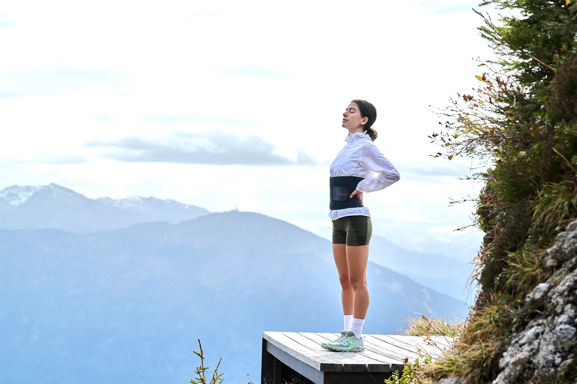 Frau steht mit geschlossenen Augen vor einem Bergpanorama, stützt die Hände in die Hüfte und trägt dabei einen ARTZT neuro Atemgürtel.