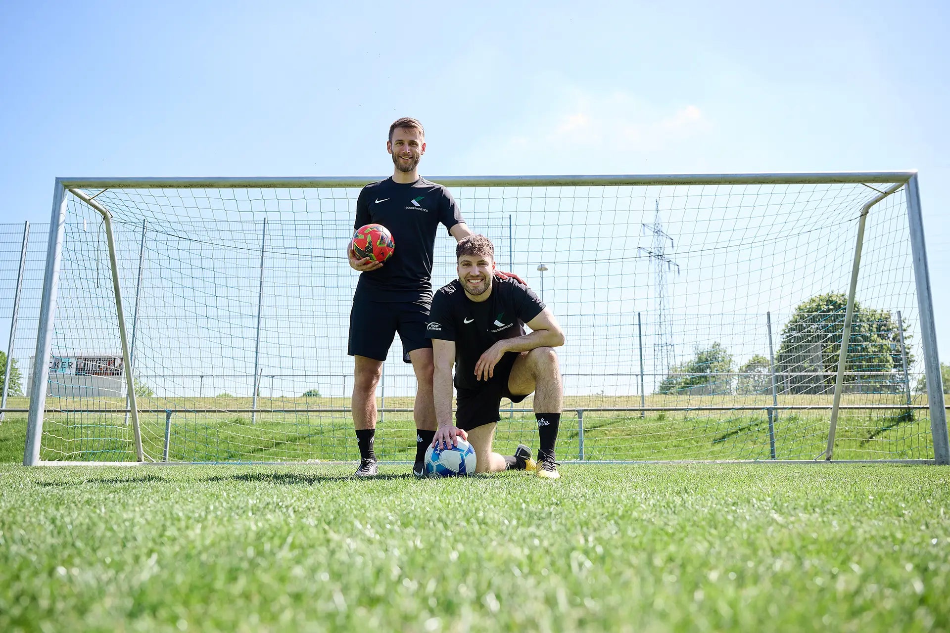 Die beiden Inhaber von Soccerkinetics stehen in Fußballtrikots auf Rasen vor einem Fußballtor. Der linke steht aufrecht und hält einen Fußball in der rechten Hand, der rechte hockt auf dem Boden und stützt sich mit der rechten Hand auf einem zweiten Fußball ab.
