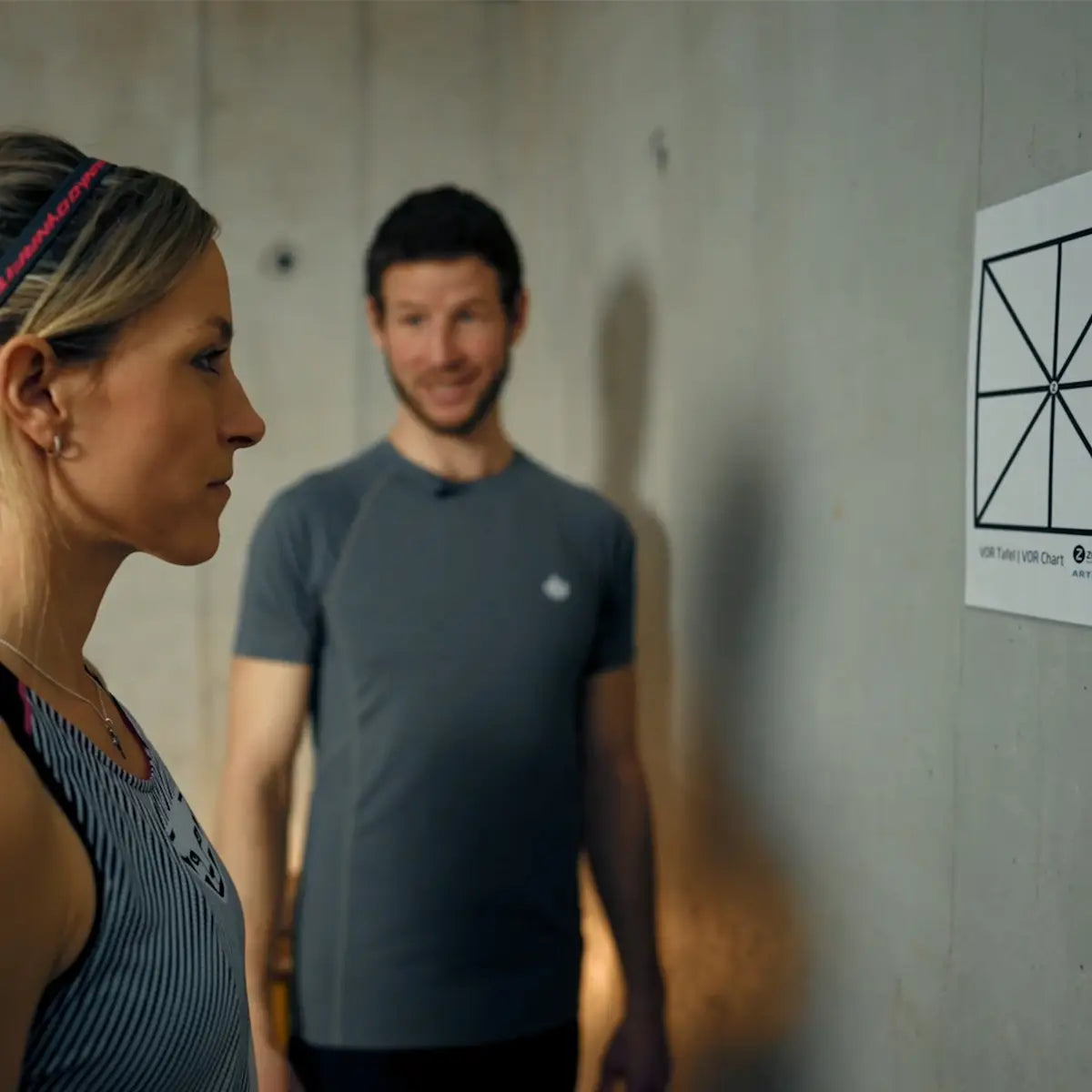 Extremsportlerin Gela Allmann schaut auf eine ARTZT neuro Sehtafel, die an einer Wand hängt, beobachtet von einem Mann im Hintergrund.