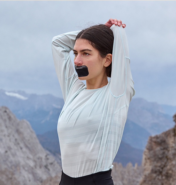 Eine Frau steht vor einem Bergpanorama und dehnt ihren linken Oberarm hinter dem Kopf, während sie ein schwarzes ARTZT neuro Mouth Tape auf dem Mund trägt.