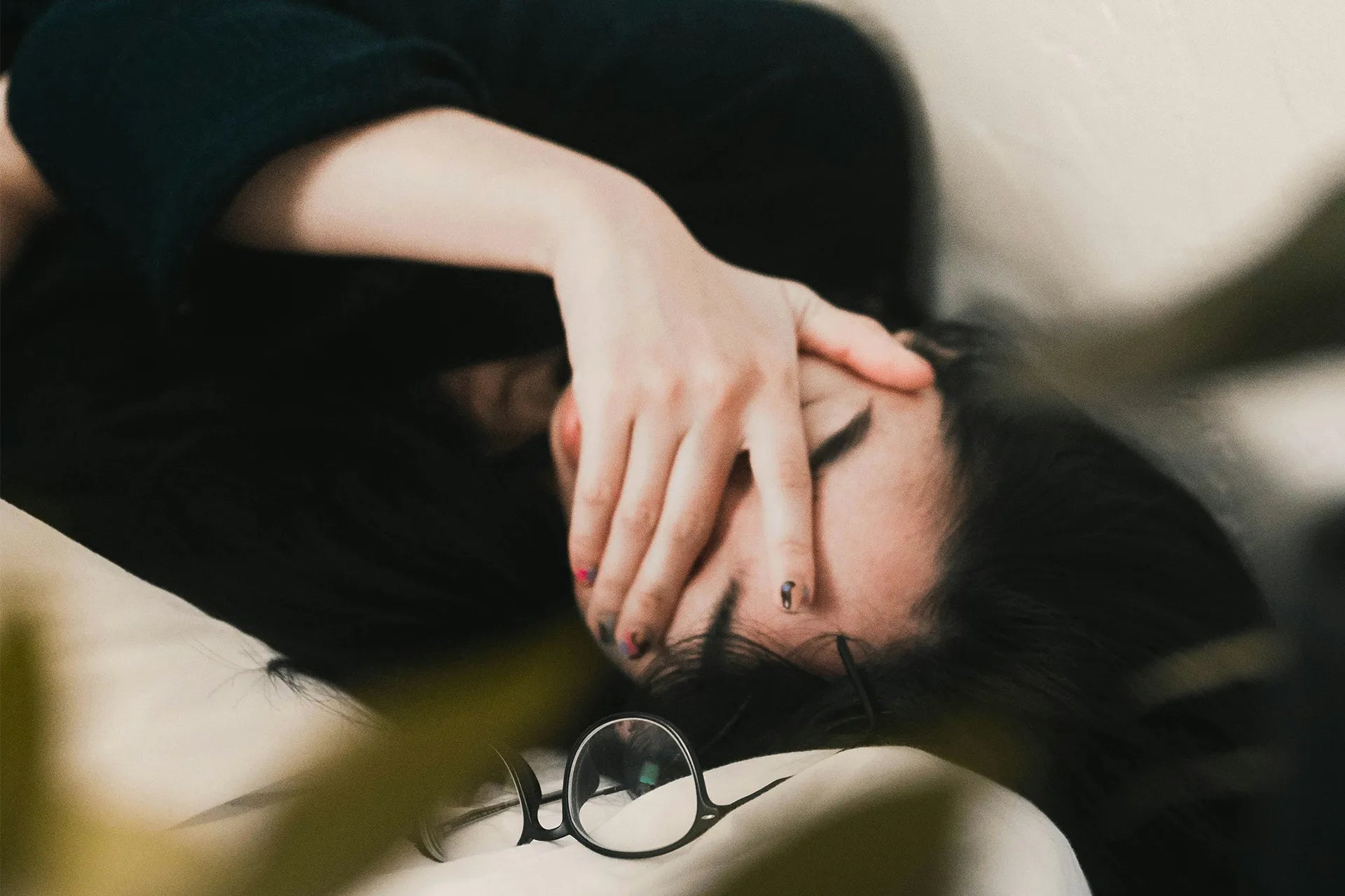 Person liegt seitlich oder leicht zurückgelehnt, bedeckt mit einer Hand das Gesicht. Eine Brille liegt neben dem Kopf auf einer hellen Unterlage, unscharfe Natur- oder Innenraumumgebung im Hintergrund. | ARTZT neuro Atemtrainer