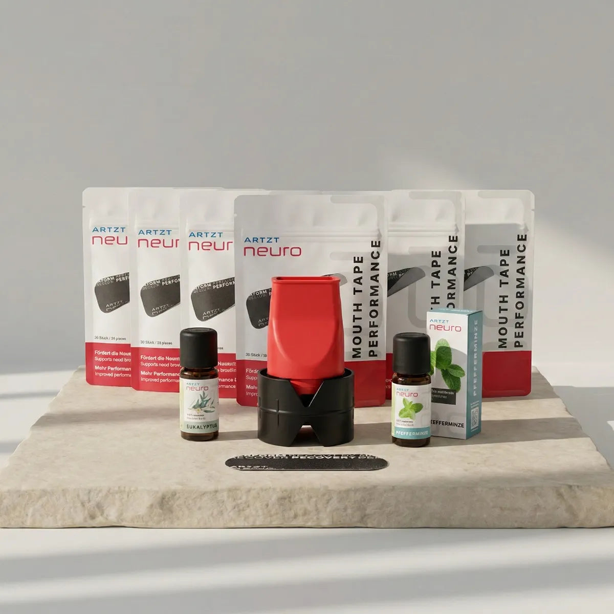 Produktaufnahme des ARTZT neuro Performance Breathing Set mit sechs Packungen Mouth Tape Performance, zwei ätherischen Ölen (Eukalyptus und Pfefferminze), einem roten Atemtrainer mit schwarzem Standfuß sowie einem einzelnen Mouth Tape auf einer hellen Steinplatte vor neutralem Hintergrund. SET-1026, SET-1027 #mengenrabatt_6 monate