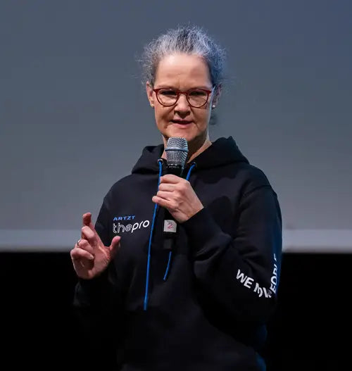 Eine Frau steht vor einem grauen Hintergrund und spricht in ein Mikrofon.