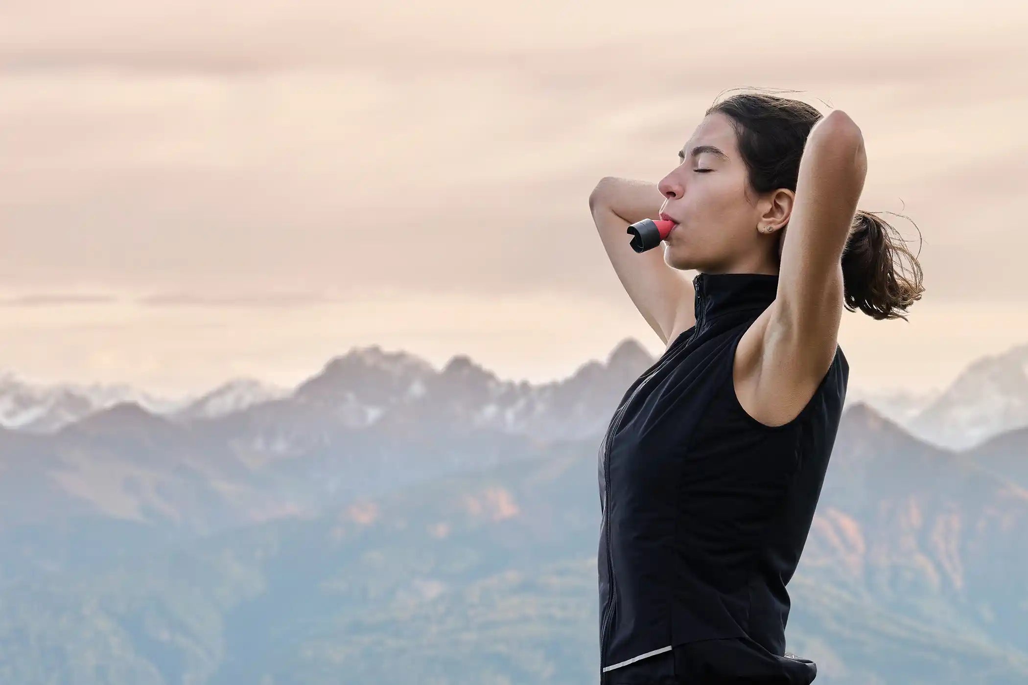 Eine Frau steht vor einem Bergpanorama mit geschlossenen Augen und betreibt Atemtraining mit dem ARTZT neuro Atemtrainer