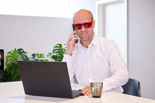 Mann sitzt am Schreibtisch vor einem Laptop mit Handy am Ohr und trägt dabei eine rote ARTZT neuro Farbbrille.