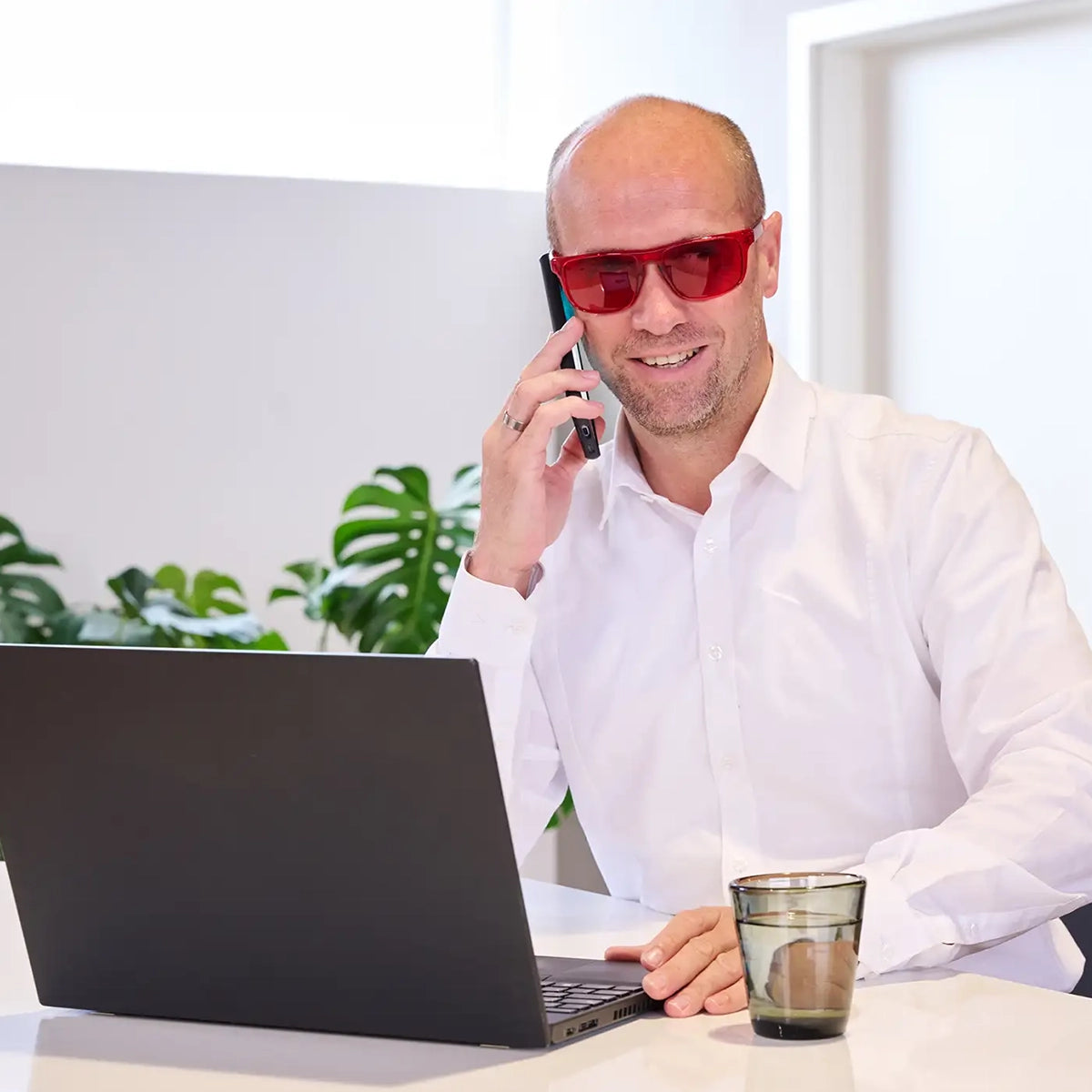 Ein Mann sitzt in einem hellen Raum an einem Scheibtisch vor einem Laptop und hält ein Smartphone an sein rechtes Ohr, während er eine rote ARTZT neuro Farbbrille trägt.