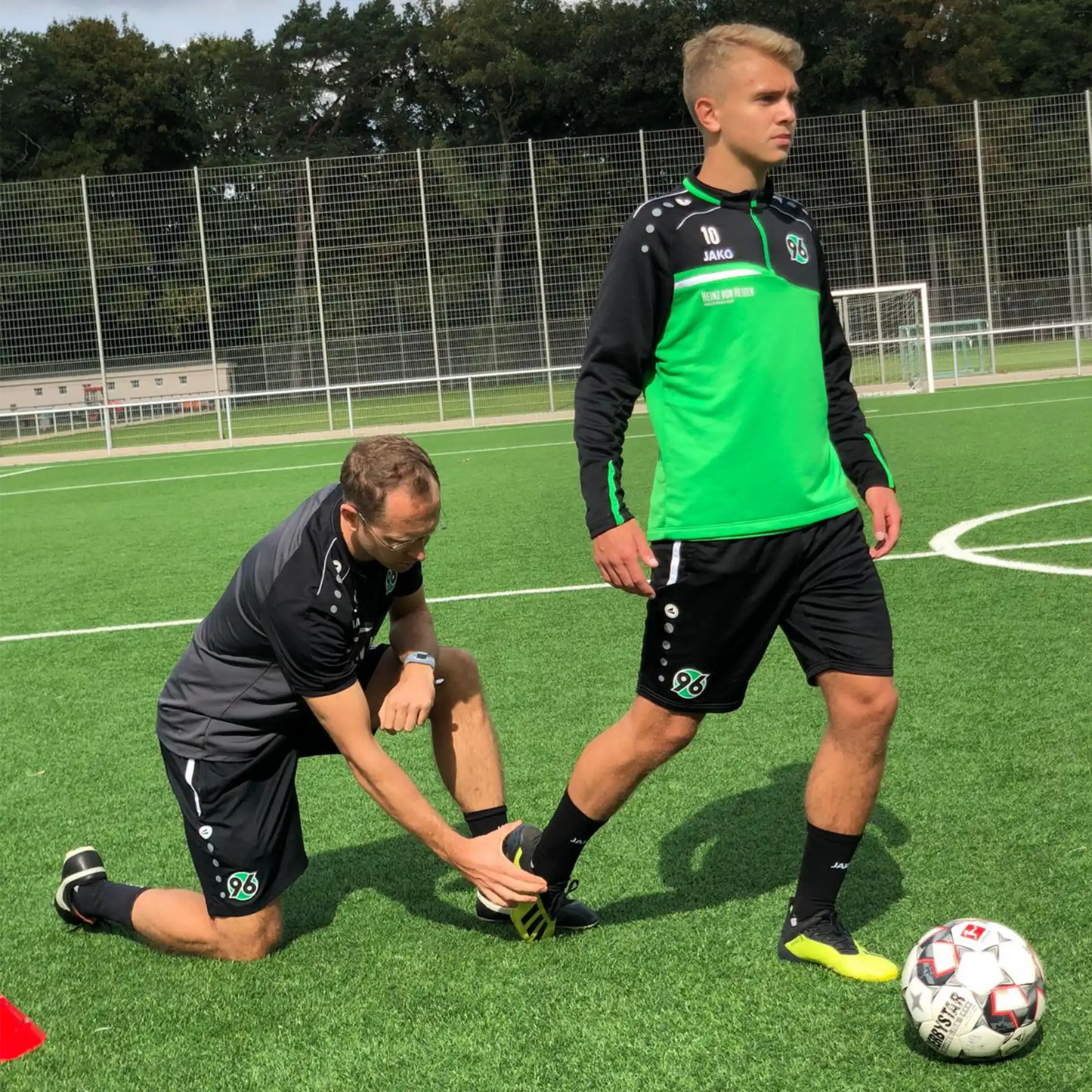 Ein Fußballspieler steht auf dem Spielfeld, während ein Fußballtrainer seine Fußhaltung kontrolliert.