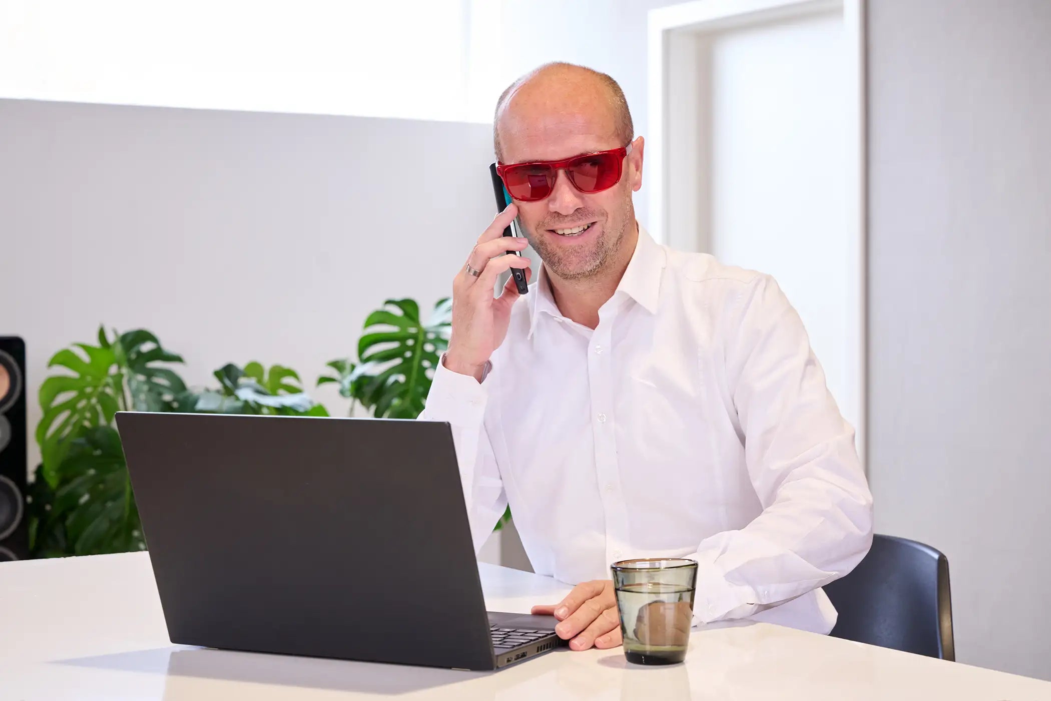 Mann sitzt am Schreibtisch vor einem Laptop und trägt eine rote ARTZT neuro Farbbrille, während er mit einem Handy telefoniert.