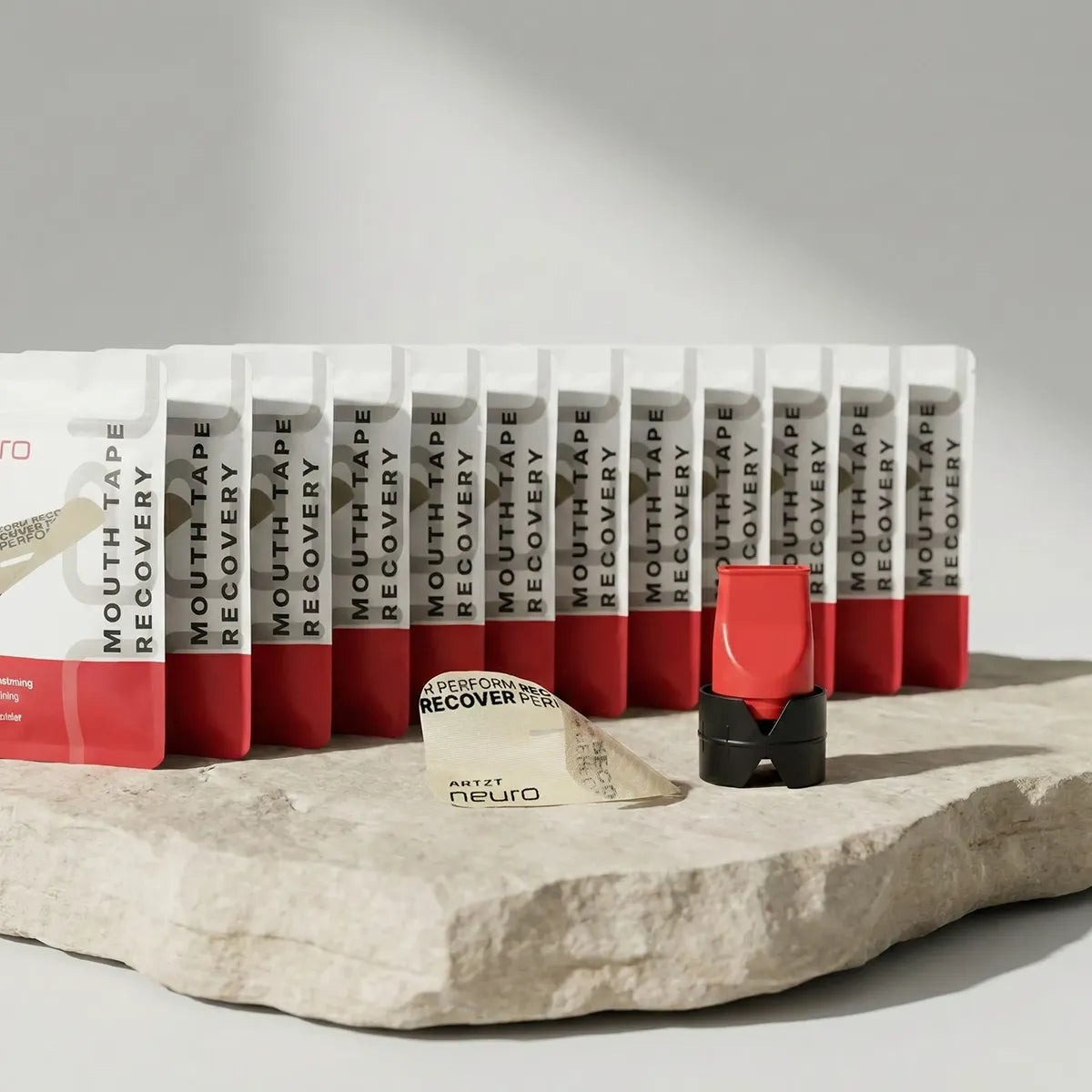 Produktaufnahme des ARTZT neuro Recovery Breathing Set mit mehreren hintereinander aufgestellten Packungen Mouth Tape Recovery, einem roten Atemtrainer mit schwarzem Standfuß sowie einem einzelnen Mouth Tape auf einer strukturierten Steinplatte vor hellem Hintergrund. SET-1021 #mengenrabatt_12 monate